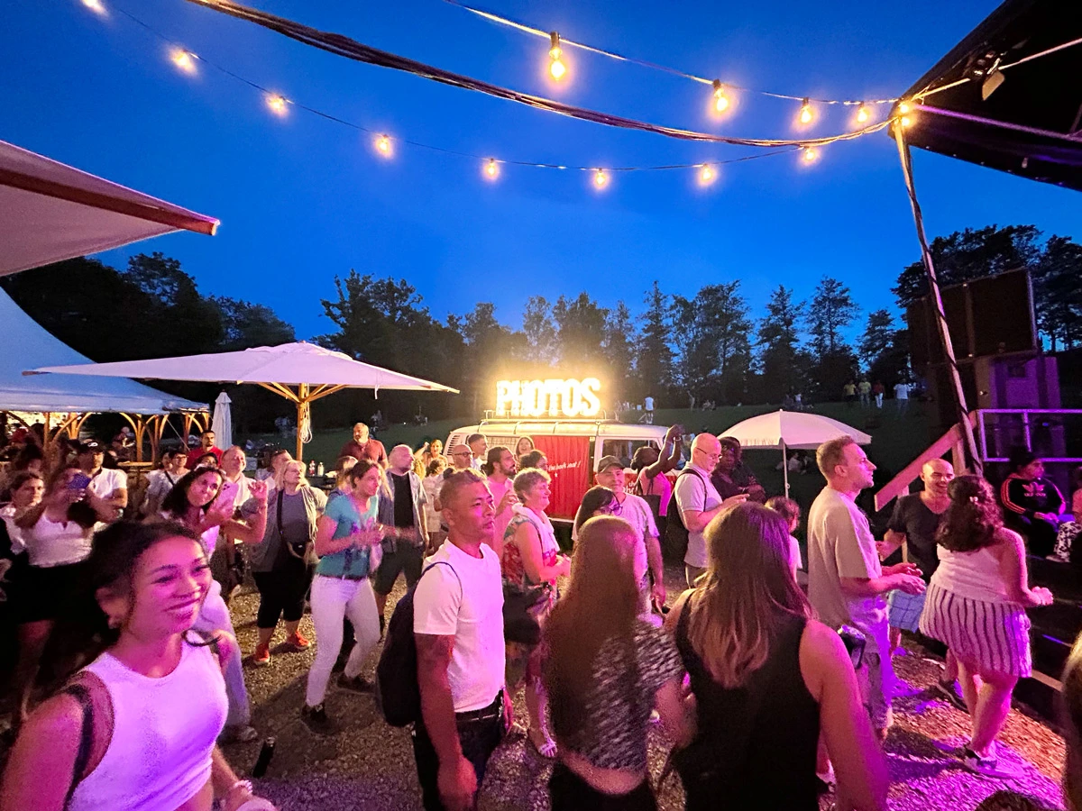 Menschen tanzen am Flughafenfest Zürich 2023 vor dem beleuchteten Photobus, festliche Lichter und sommerliche Abendstimmung.