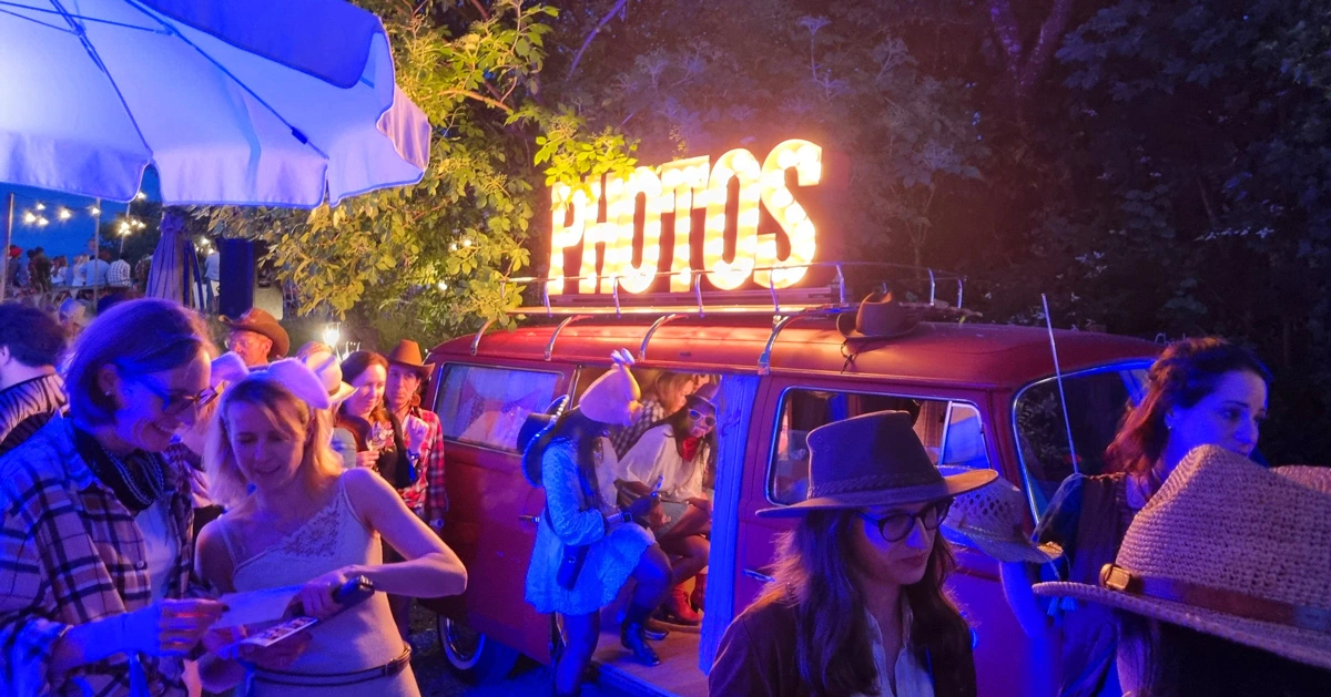 Glückliche Mitarbeiter*innen stehen Schlange an einem roten Fotobus mit leuchtender Photos-Schrift bei einem Firmen-Event am Abend.