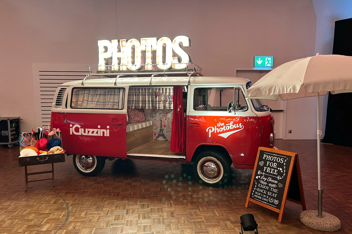 Mitarbeiter*innen stehen gut gelaunt Schlange vor einem leuchtenden roten Photobus bei einer Abendveranstaltung.