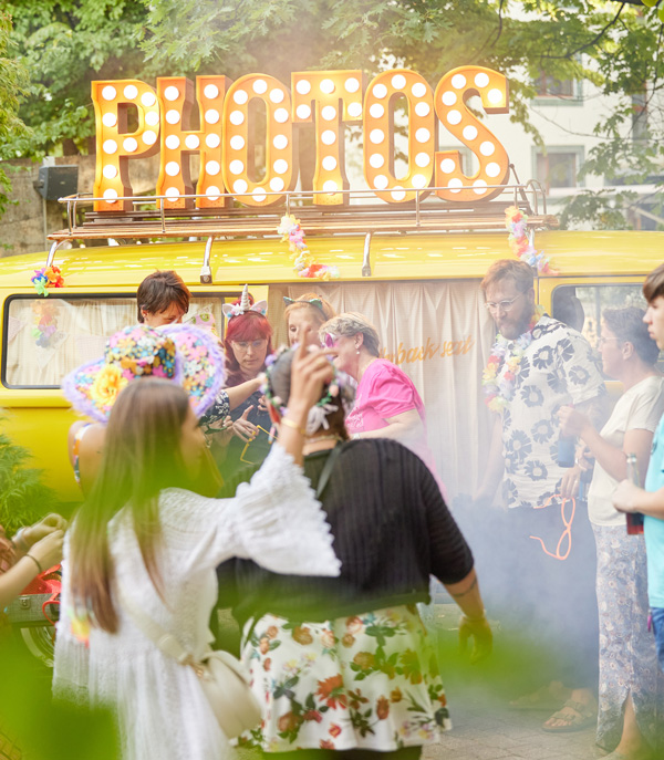 Fröhliche Partygäste posieren und lachen rund um einen gelben VW Fotobus mit beleuchtetem 'PHOTOS' Schild auf einem Event im Freien.