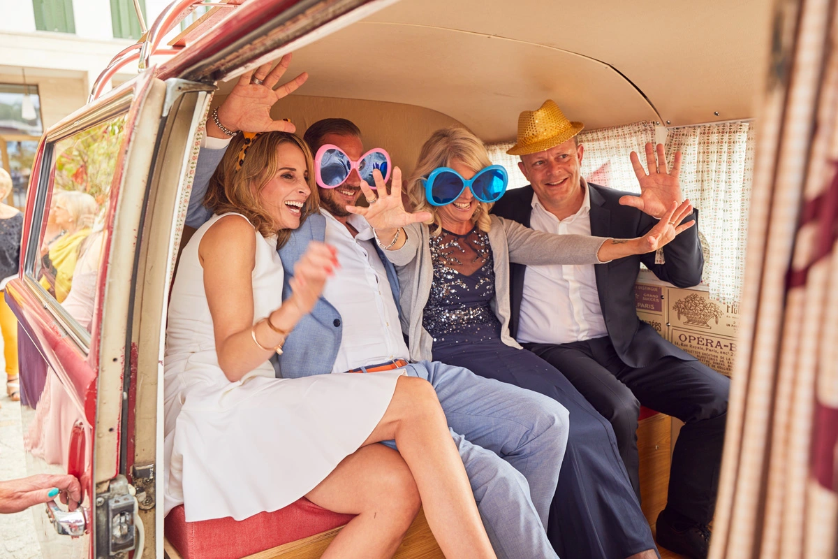 Vier lachende Kolleg*innen mit bunten XXL-Brillen und Partyhüten posieren gemeinsam auf der Rückbank eines Photobusses.