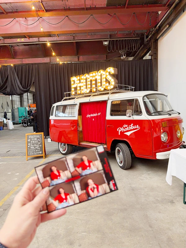 Eine Hand hält einen frisch gedruckten Fotostreifen vor dem roten VW Photobus in einer Eventlocation.