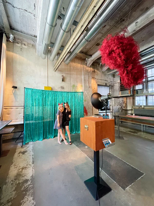 Zwei Frauen mit Requisiten posieren vor grünem Glitzerhintergrund an der Stand-Fotobox von The Photobus in einer Industrielocation