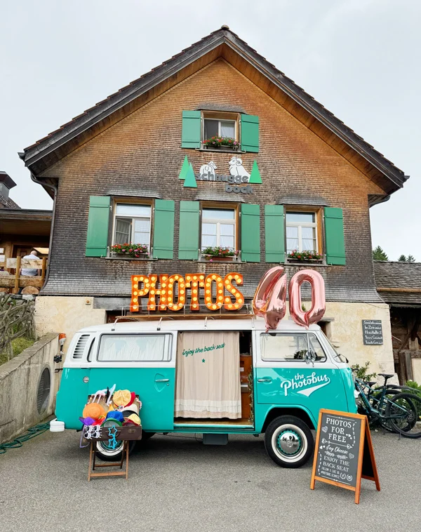 Mintgrüner VW Photobus mit Leuchtschrift PHOTOS und rosagoldenem Luftballon 40 für einen 40. Geburtstag in der Schweiz, mit Requisiten-Koffer und Kreidetafel vor rustikalem Schweizer Bauernhaus