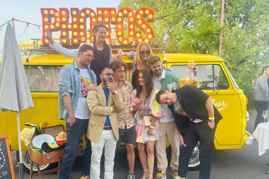 Gruppe von Mitarbeitern feiert ausgelassen vor dem gelben VW Photobus bei einem Firmenevent im Freien in der Schweiz mit leuchtendem PHOTOS-Schriftzug
