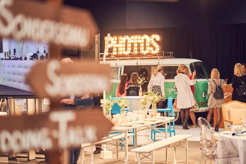 Grüner Photobus VW-Bus mit leuchtendem PHOTOS-Schild an der Hochzeitsmesse in den Olma Messehallen St. Gallen, elegante Tischdekoration im Vordergrund