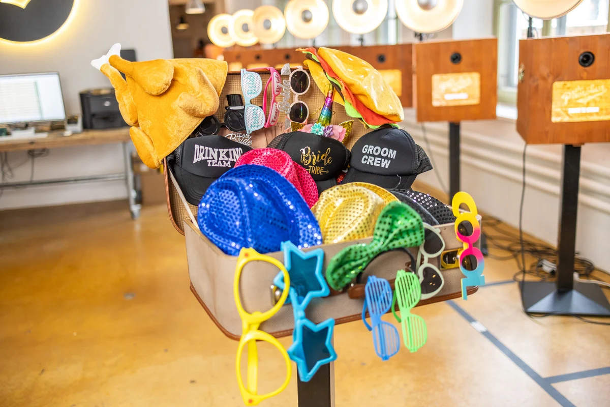 Aufgeklappter Requisiten-Koffer mit bunten Brillen, Hüten und Accessoires vor dem Photobus-Logo und mehreren Stand-Fotoboxen im Hintergrund