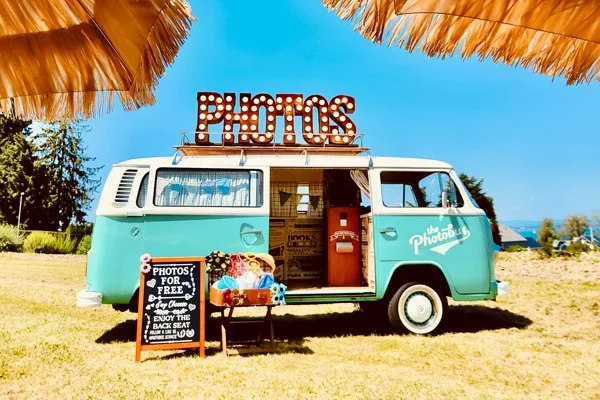 Türkiser Photobus VW-Bus mit leuchtendem PHOTOS-Schild und Requisiten aufgebaut auf einer Sommerwiese bei strahlendem Sonnenschein
