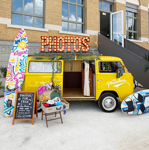 Gelber Photobus mit PHOTOS-Leuchtschrift, Surfbrettern und Requisiten vor einem Firmengebäude an einem Sommerfest